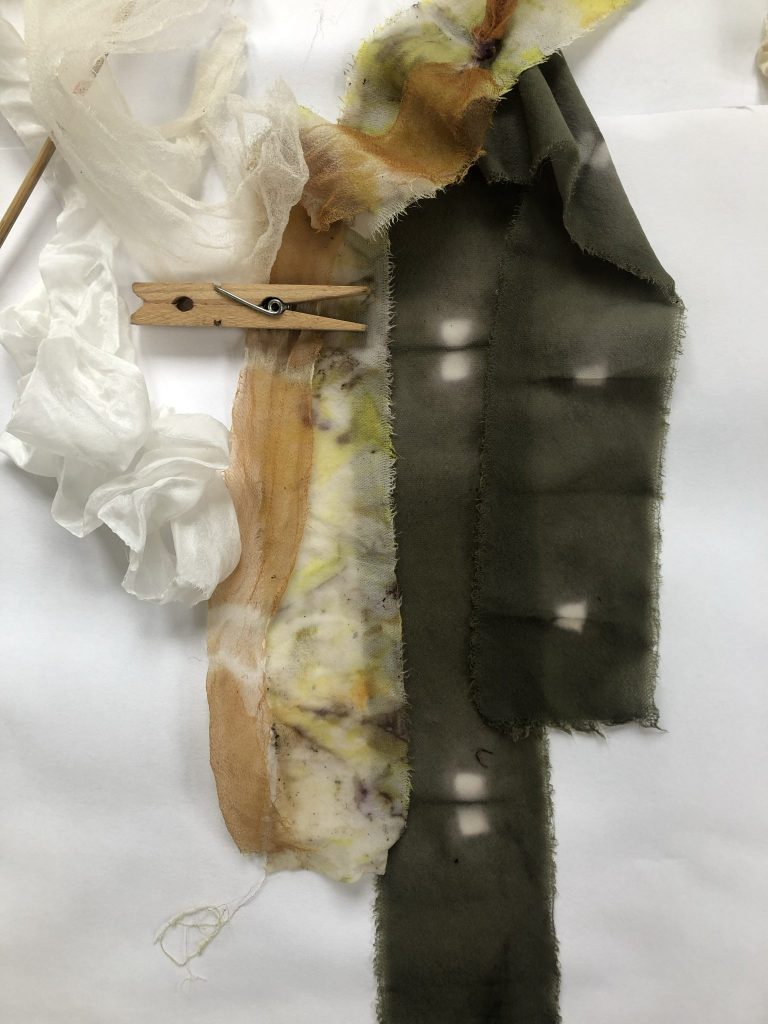 Strips of dyed fabric in earthy tones clipped together with a wooden peg, showing textures and colour variations.