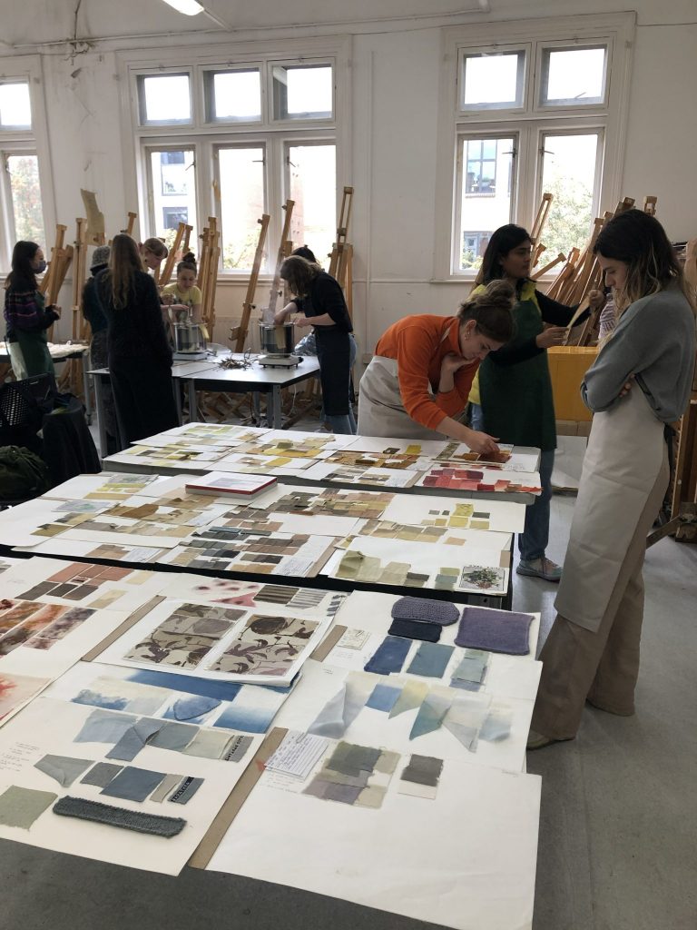 Students in a classroom working with dye baths and laying out fabric swatches on large tables covered in colourful samples.