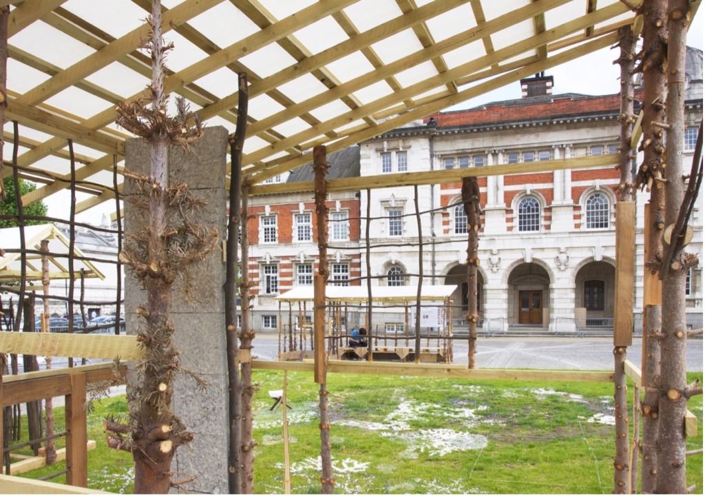 Wooden framework structure with tree trunks and branches forming a pavilion on the Chelsea College of Arts parade ground.