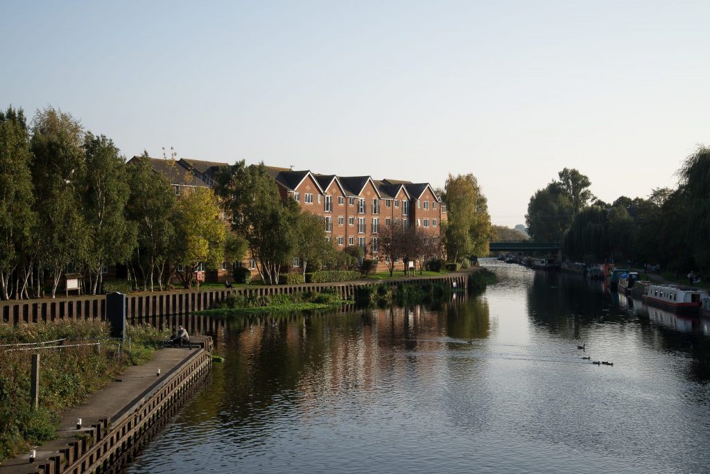 Calm river scene with red-brick houses, trees, and reflections in the water under a clear sky.