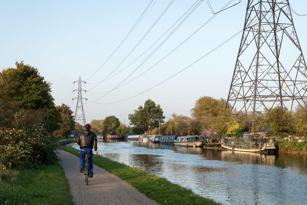 Person cycling along a canal path lined with boats and overlooked by electricity pylons.