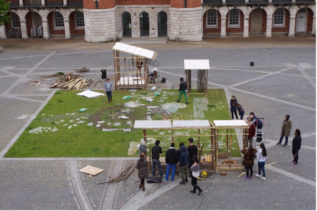 Aerial view of students and staff assembling garden structures and arranging natural materials on a grass square.