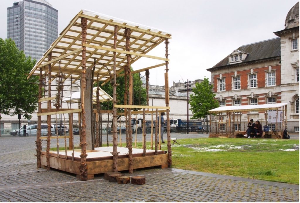 Pavilion-like wooden structures built from tree trunks and reclaimed materials in the open parade ground.