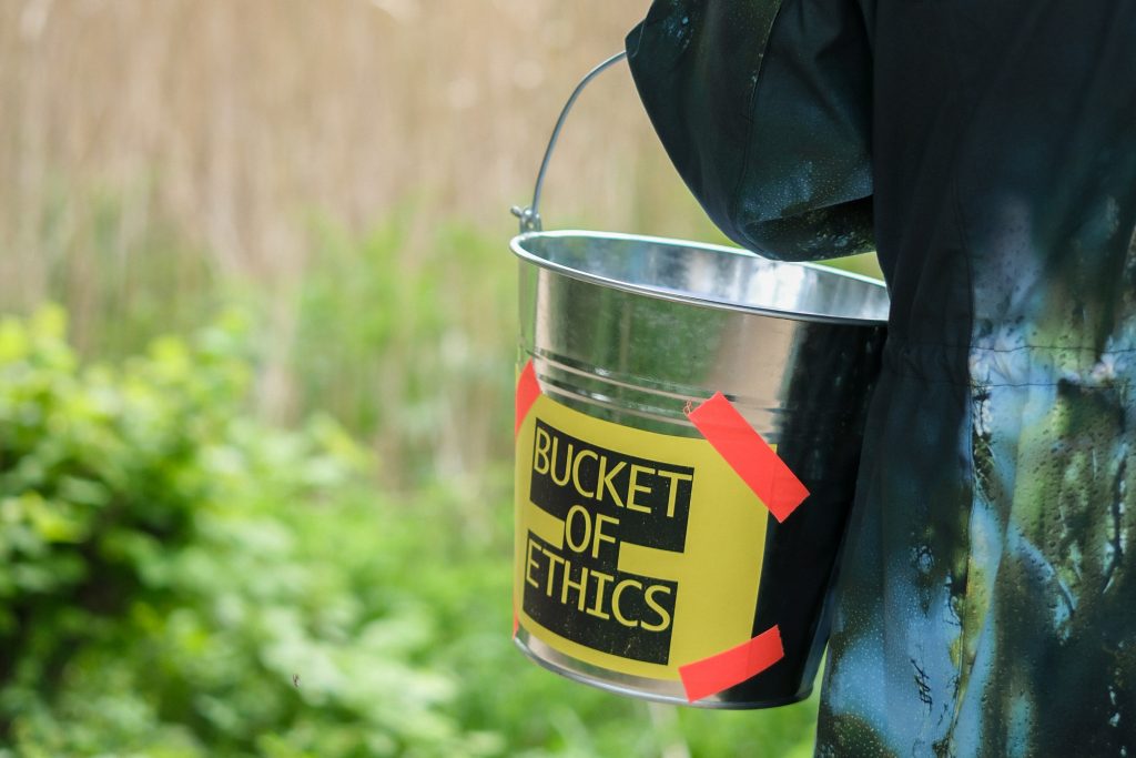 Metal bucket labeled “Bucket of Ethics” taped with red corners, carried outdoors.