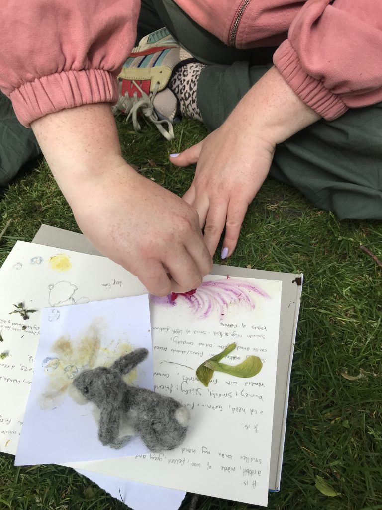 Close-up of hands arranging natural objects and drawings on paper, with a small felt rabbit placed on top.