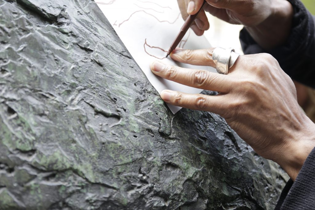 Close-up of hands making a rubbing with a brown pen on paper pressed against a rough green stone surface.