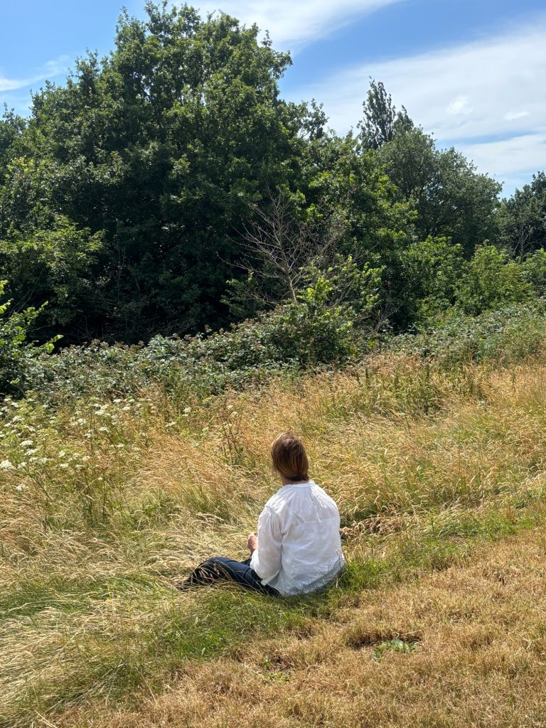 Participant sitting quietly in tall grass on Hampstead Heath, reflecting and observing the natural environment.