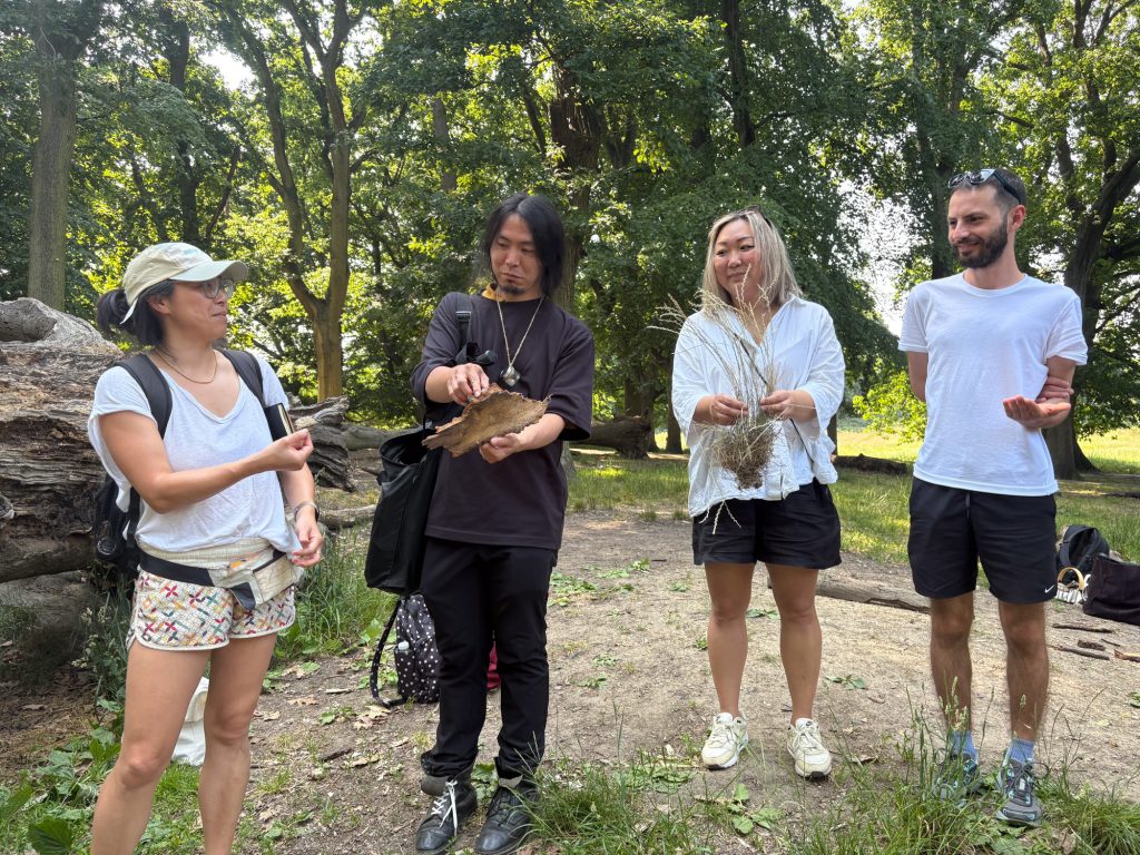 Four participants standing together outdoors, each holding different natural materials such as bark, grass, and twigs, sharing observations.